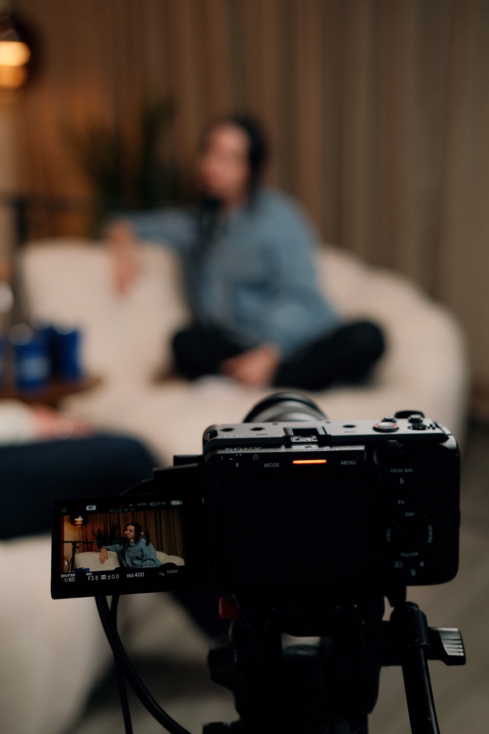 A camera on a tripod filming a guest sitting on a sofa at a podcast mic